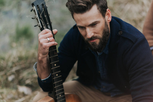 白人髭の男が湖の近くでギターを弾いてる。ハンサムなスタイリッシュなヒップスター. - 写真・画像