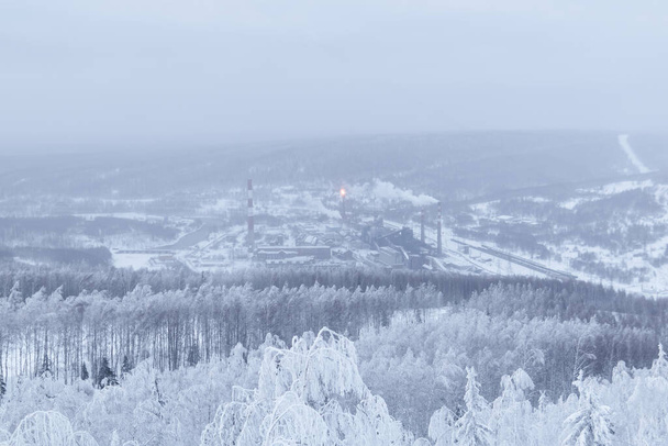 winter landscape - a distant metallurgical plant in a valley in the middle of snow-covered forests in a frosty haze - Photo, Image