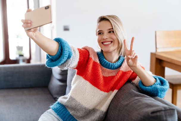 Happy young woman taking a selfie while sitting on a couch at home - Φωτογραφία, εικόνα