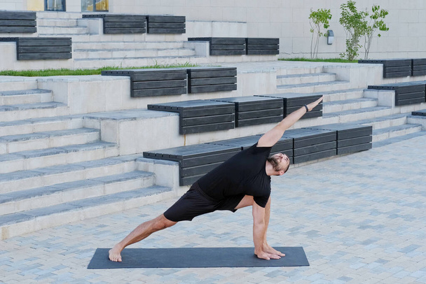 Mann Sportler in schwarzer Sportbekleidung praktiziert Yoga-Übungen in Kriegerpose im Freien - Foto, Bild