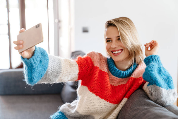 Happy young woman taking a selfie while sitting on a couch at home - Photo, Image