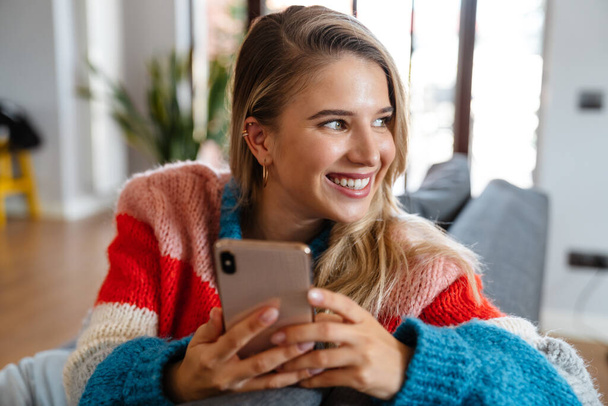 Attractive smiling young woman using mobile phone while ralaxing on couch at home - Fotografie, Obrázek