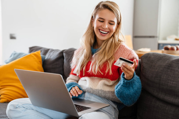 Lovely young attractive woman relaxing on a couch in the living room, using laptop computer, showing credit card, celebrating - Fotoğraf, Görsel