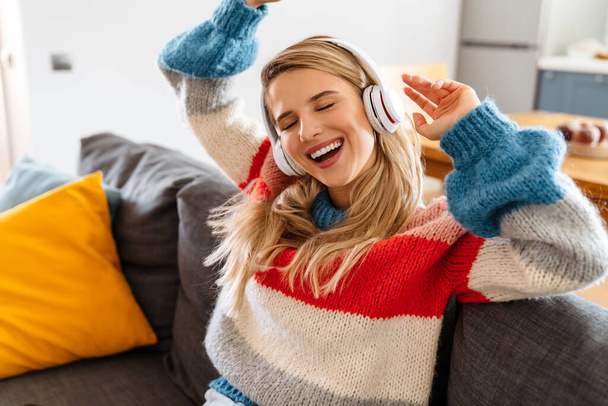 Happy young woman sitting on sofa listening to music with headphones, dancing - Photo, Image