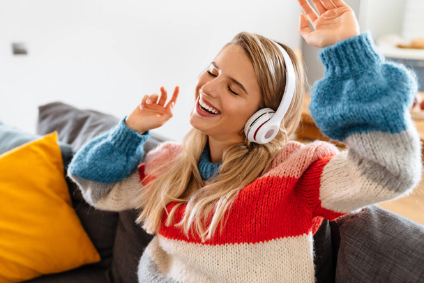 Happy young woman sitting on sofa listening to music with headphones, dancing - Фото, зображення
