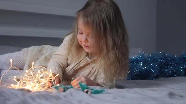 ładna dziewczynka bawiąca się małymi choinkami i girlandą leżącą na łóżku w szarej sypialni. Nowy rok, dzieciństwo, córka. Nagrania w Full HD - Materiał filmowy, wideo