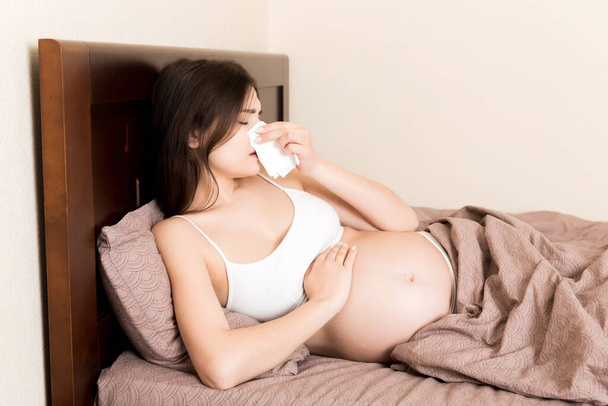 Portrait of young unhappy pregnant woman leaning on pillows on bed and blowing her nose into tissue. Future mom caught cold and sneezing. - Фото, изображение