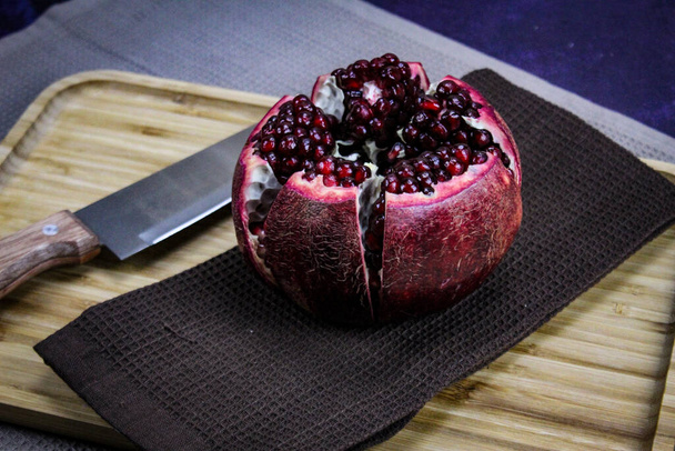 Egy nyitott gránátalma gyümölcs, amely öt részre van osztva, amelyeket egy szalvétán / szöveten tartanak egy fa tálcán, kék alapon. Nagy piros érett gránátalma, mellette egy késsel.. - Fotó, kép