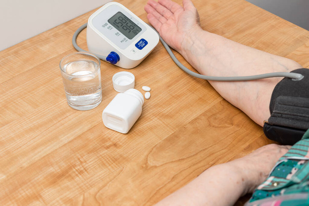 Žena má hypertenzi, měří krevní tlak na ruce s digitálním tonometrem, hypertenzní léky, léky na léčbu a sklenice vody na stole. - Fotografie, Obrázek