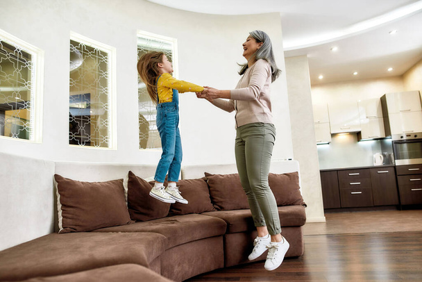 Fun time. Happy granddaughter jumping on a couch while having fun with her cheerful grandmother at home - Φωτογραφία, εικόνα
