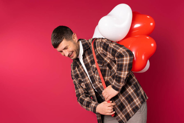 Young cute man carries a bunch of heart-shaped balloons on his back. St.Valentines Day on red background and side space. High quality photo - Foto, Imagem
