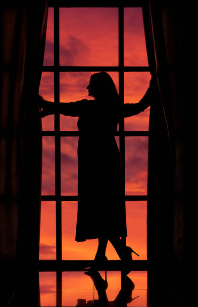 gyönyörű sziluett egy nő egy hosszú peignoir egy nagy ablak hátterében egy narancssárga naplemente ég - Fotó, kép
