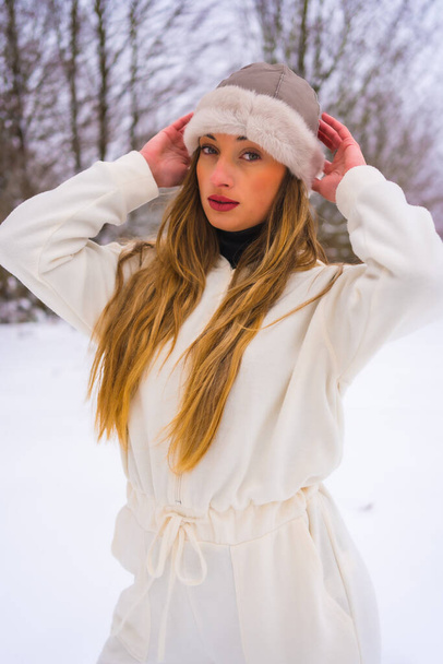 Winter lifestyle, portrait caucasian blonde with white winter outfit and woolen hat in the snow next to snowy pine trees, vacation in nature - Foto, immagini