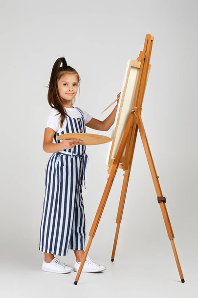 Portrait of beautiful little girl holding a wooden art palette and brush on studio background - Fotoğraf, Görsel