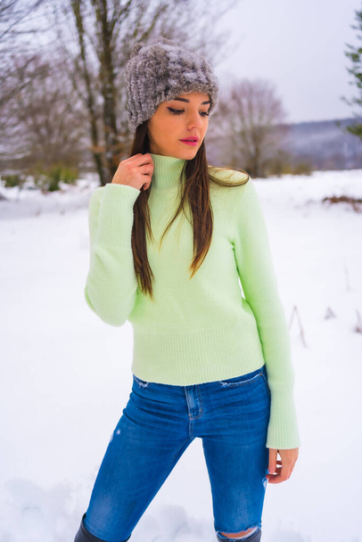 Winter lifestyle, portrait young brunette caucasian with green outfit and woolen hat enjoying the snow next to snowy pine trees, vacations in nature - Foto, immagini