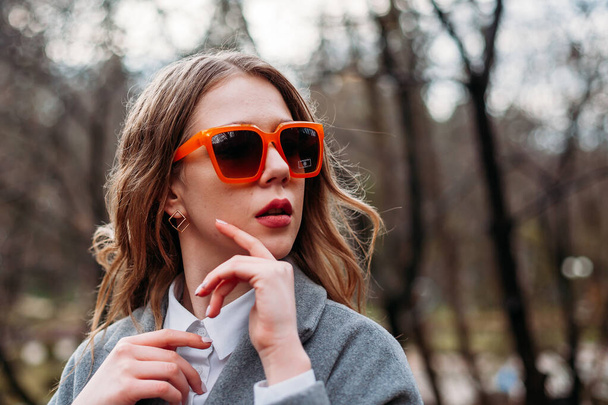 young woman in a gray coat against the background of a gray autumn forest in bright contrasting orange glasses. bright mood concept - Foto, Imagem