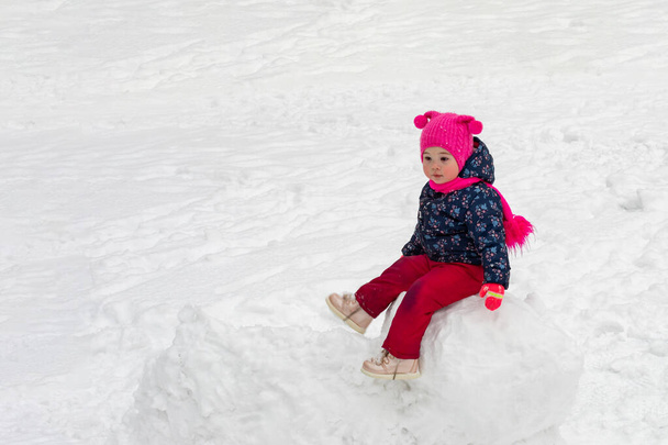 Little caucasian girl in a pink hat sitting on a snow ball, winter fun making a snowman - Photo, Image