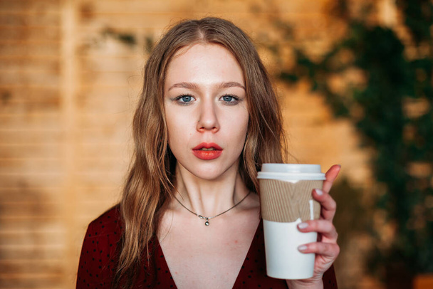Young blonde woman holding a paper cup of hot coffee of takeaway in her hands indoors in cafe and looking in camera. copyspace. Coffee shop advertisement. Place for your text, concept business drinks. - Foto, Bild