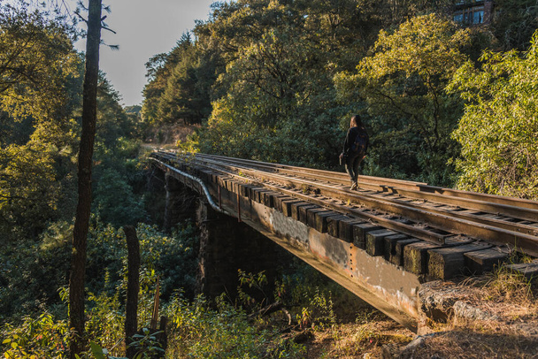 Cserkészlány sétál és pihen a vasúti sínek mentén, és egy híd az erdő közepén, fekete ruhában, kék hátizsákkal, fekete kalapban és hátizsákkal 12 - Fotó, kép
