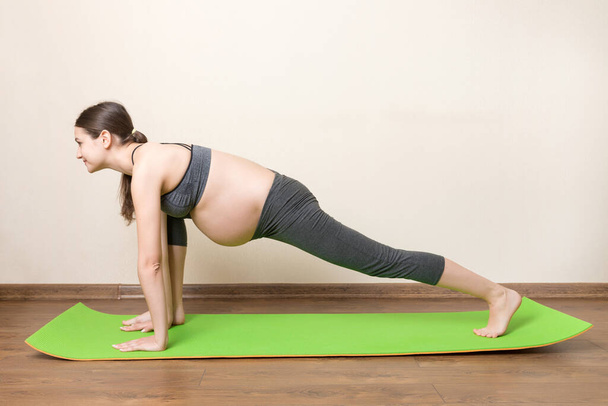 mooie jonge zwangere vrouw zitten op mat in yoga pose bij het huis. Zwangerschap Yoga en Fitness concept op coronavirus tijd. - Foto, afbeelding