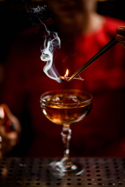 Close-up view of glass with drink over which bartender holds smoldering plant - Φωτογραφία, εικόνα
