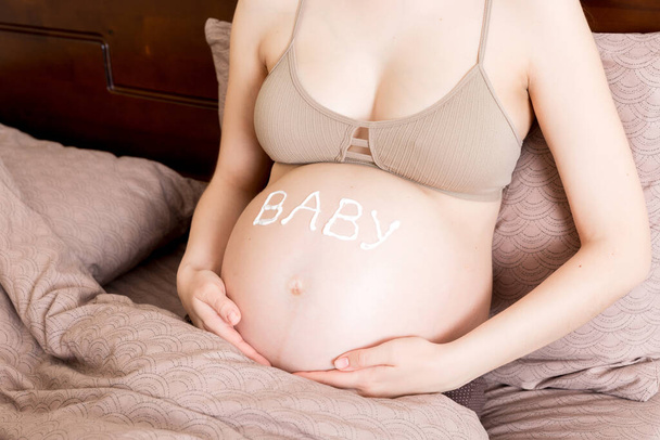 Egy terhes lány otthon ül az ágyon, és egy nyúlásgátló krémet kent a hasára. Terhesség, anyaság, felkészülés és elvárás koncepció.a szó baba a hason. - Fotó, kép