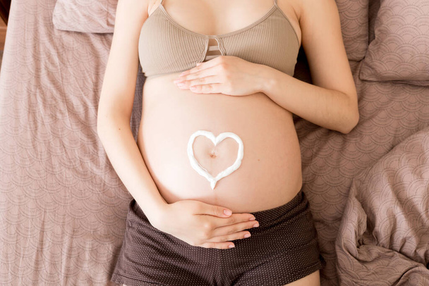 a pregnant girl sits at home on the bed and smears heart an anti-stretch mark cream on her stomach. Pregnancy, motherhood, preparation and expectation concept. - Photo, Image