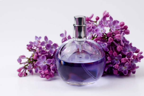 Bottiglia di profumo di vetro in composizione con ramo aromatico di fiori lilla su sfondo bianco con spazio per copiare il testo. Modello di biglietto di auguri - Foto, immagini