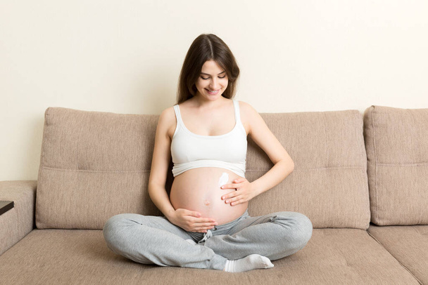 raskaana oleva tyttö istuu kotona sängyllä ja tahraa anti-venytys merkki kerma hänen vatsaan. Raskaus, äitiys, valmistelu ja odotukset käsite. - Valokuva, kuva