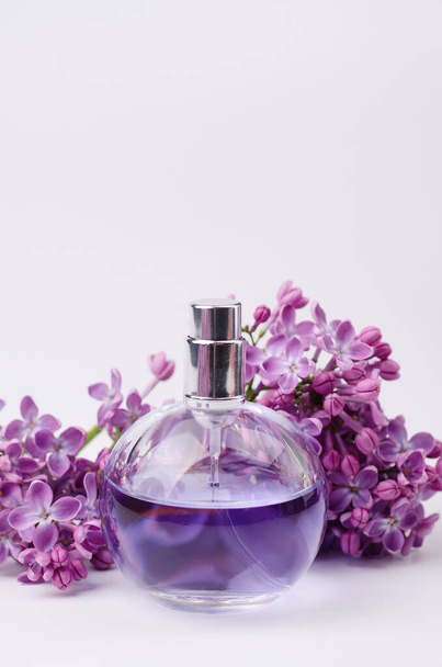 Bottiglia di profumo di vetro in composizione con ramo aromatico di fiori lilla su sfondo bianco con spazio per copiare il testo. Modello di biglietto di auguri - Foto, immagini