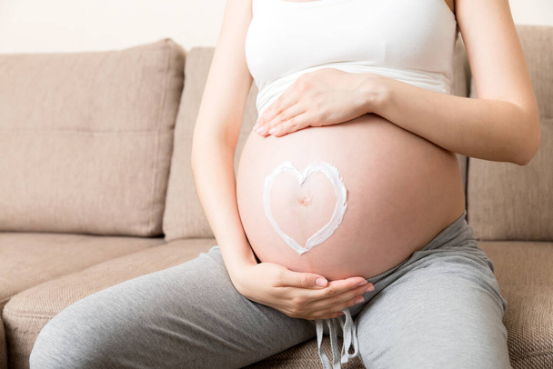 Μια έγκυος κάθεται στο κρεβάτι και πασαλείβει την καρδιά της με κρέμα κατά των ραγάδων στο στομάχι της. Εγκυμοσύνη, μητρότητα, προετοιμασία και προσδοκία. - Φωτογραφία, εικόνα