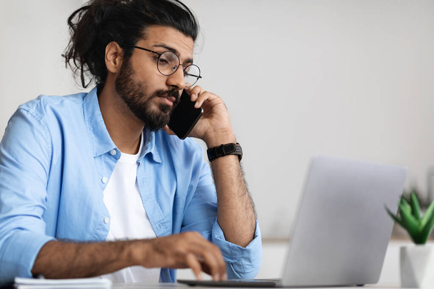 Занятой молодой арабский бизнесмен разговаривает по мобильному телефону и работает за портативным компьютером - Фото, изображение