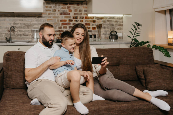 Ένας πατέρας, ένας γιος και μια νεαρή μητέρα βλέπουν ένα βίντεο σε ένα smartphone στον καναπέ. Μια μαμά δείχνει στην οικογένειά της κάτι ενδιαφέρον στην οθόνη του κινητού της το βράδυ στο σπίτι.. - Φωτογραφία, εικόνα