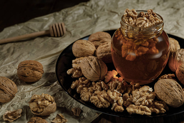 Honey and walnuts in a glass jar on a black plate. Background of crumpled paper and honey sticks. - Fotografie, Obrázek