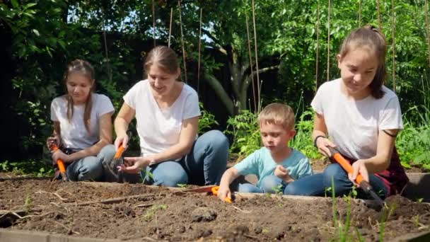 Los niños ayudan a la madre a trabajar en el jardín y plantar verduras en la cama de jardín en el patio trasero. Concepto de trabajo en equipo y trabajo en familia - Imágenes, Vídeo