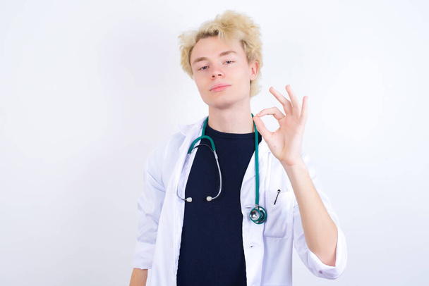 Jsem rád, že atraktivní mladý pohledný běloch doktor v bílém plášti ukazuje ok znamení s rukou jako vyjádření souhlasu, má veselý výraz, je optimistický. - Fotografie, Obrázek