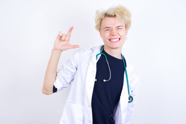 junge hübsche kaukasische Arzt trägt weißen Mantel lächelnd und gestikulierend mit der Hand klein, Maß Symbol. - Foto, Bild