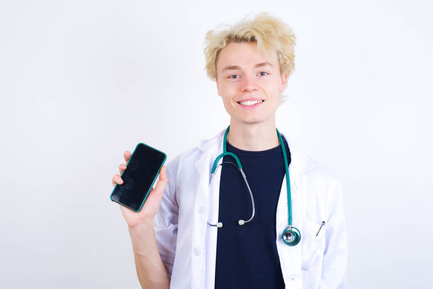 Χαμογελαστός νεαρός ξανθός γιατρός με λευκό παλτό και στηθοσκόπιο που δείχνει άδεια τηλεφωνική οθόνη. Έννοια διαφήμισης και επικοινωνίας. - Φωτογραφία, εικόνα