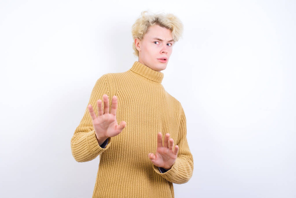 Afraid handsome Caucasian blond man standing against white background makes terrified expression and stop gesture with both hands saying: Stay there. Panic concept. - Fotografie, Obrázek