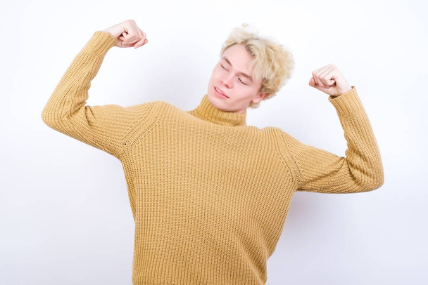 Waist up shot of handsome Caucasian blond man standing against white background raises arms to show muscles feels confident in victory, looks strong and independent, smiles positively at camera. Sport concept. - Photo, Image