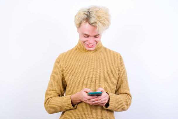 Smiling handsome Caucasian blond man standing against white background using cell phone, messaging, being happy to text with friends, looking at smartphone. Modern technologies and communication. - Photo, Image