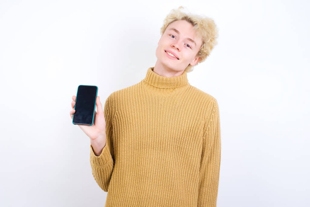 Χαμογελώντας όμορφος Καυκάσιος ξανθός άντρας στέκεται πάνω σε λευκό φόντο δείχνοντας άδεια οθόνη τηλεφώνου. Έννοια διαφήμισης και επικοινωνίας. - Φωτογραφία, εικόνα