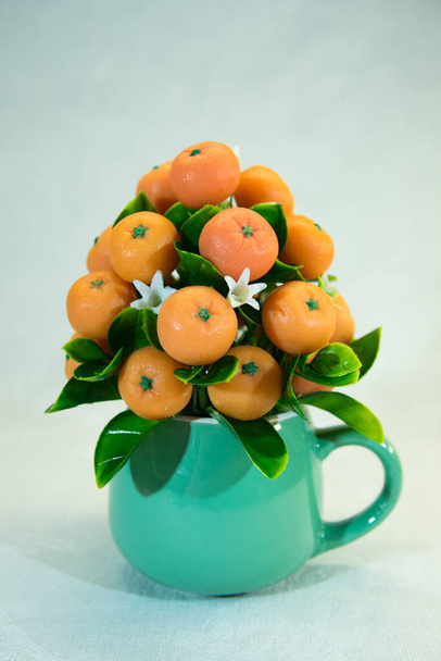 Geschenkstrauß aus leuchtend orangefarbenen Mandarinenfrüchten und weißen Blumen in einer grünen Tasse. Der Strauß ist mit grünen Blättern dekoriert. Handgemachte Mandarinen aus Seife. - Foto, Bild