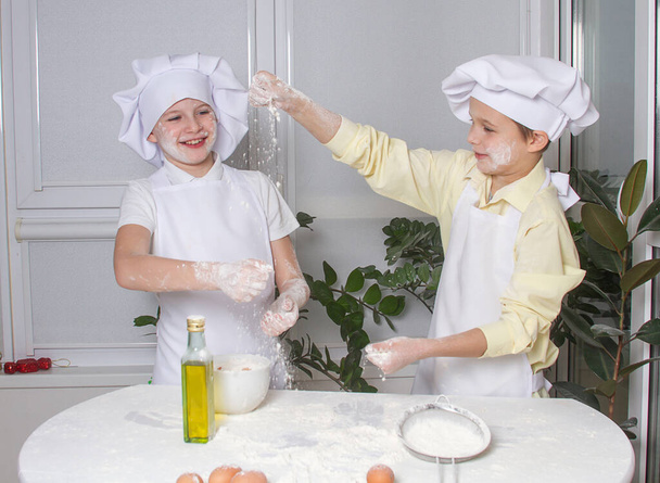 Aşçı gibi giyinmiş iki tatlı Avrupalı çocuk un, süt ve yumurta yoğuruyor, çocuklar mutfakta oynuyor, gençler hamurdan turta yapıyor.. - Fotoğraf, Görsel