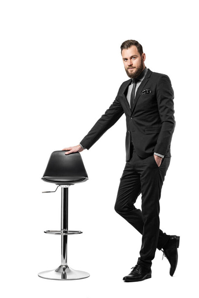 Ελκυστικός επιχειρηματίας με μαύρο κοστούμι και γραβάτα, στέκεται πάνω σε λευκό φόντο ακουμπισμένος σε καρέκλα. - Φωτογραφία, εικόνα