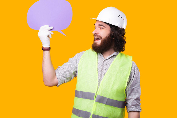 Un alegre joven arquitecto barbudo está mirando la burbuja del habla que sostiene cerca de una pared amarilla con un casco, unos guantes y un chaleco fosforescente. - Foto, Imagen