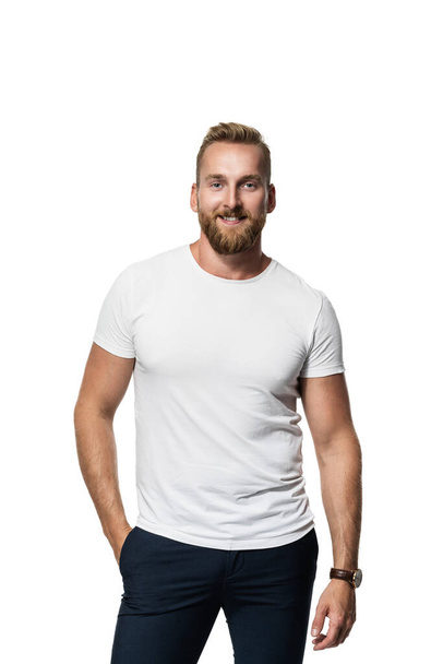 Relaxed attractive blonde bearded man standing against a white background wearing a white t-shirt, smiling towards camera. - Фото, изображение