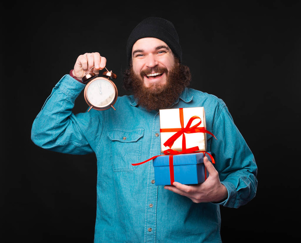 Innostuneella parrakkaalla miehellä on lahjoja ja pieni kello, joka osoittaa, että heidän aikansa on päättymässä. - Valokuva, kuva