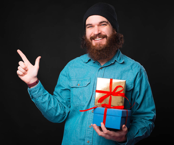 A cheerful young bearded hipster is holding some gifts and looking at the camera smiling is pointing away - Photo, Image