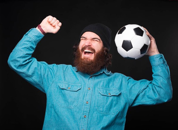 Podekscytowany młody brodaty mężczyzna trzyma piłkę nożną i krzyczy, bo wygrał przy czarnej ścianie. - Zdjęcie, obraz